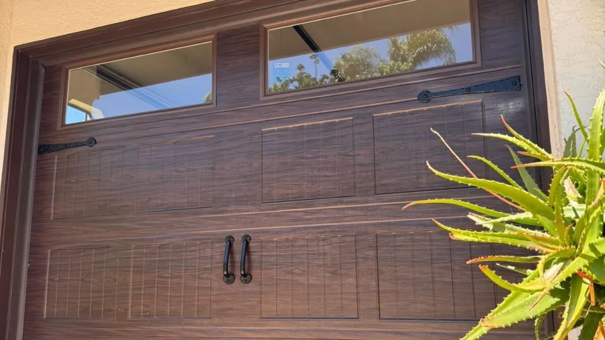 Wood-grain carriage garage door with black decorative hinges, handles, and two top windows